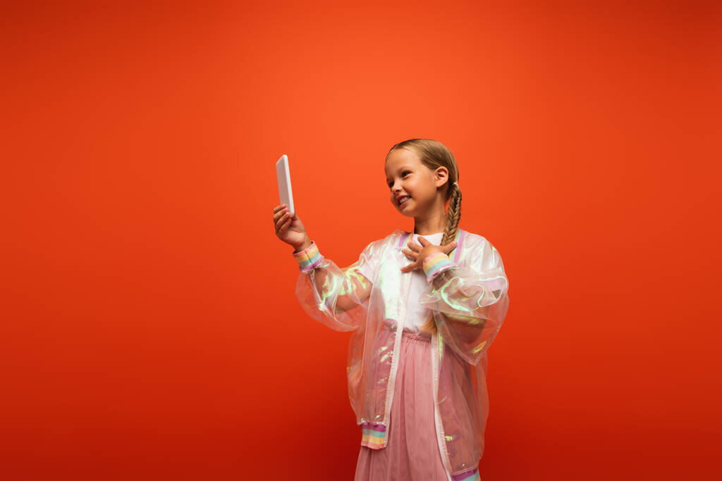 potěšená dívka v módní dešťové bundy dotýkající se hrudníku při rozhovoru během videohovoru izolované na oranžové - Fotografie, Obrázek