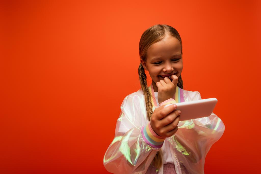 sorridente ragazza in giacca antipioggia che tiene la mano vicino alla bocca mentre guarda lo smartphone isolato su arancione - Foto, immagini