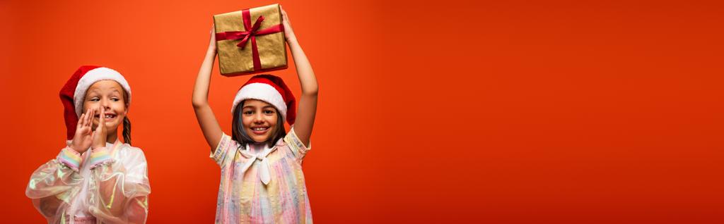 щаслива дівчина в капелюсі Санта тримає подарункову коробку в піднятих руках поруч з збудженим другом, плескаючи руками ізольовані на помаранчевому, банер
 - Фото, зображення