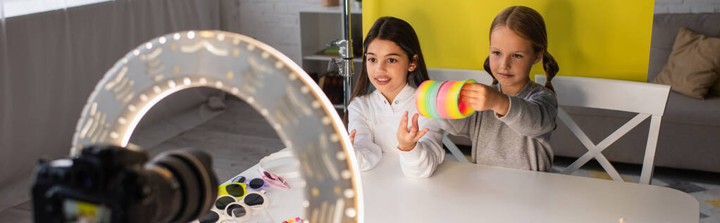 nastolatka dziewczyna pokazując spiralne zabawki w pobliżu blogger mówi przed rozmyte aparat cyfrowy w okrągłe lampy, baner - Zdjęcie, obraz