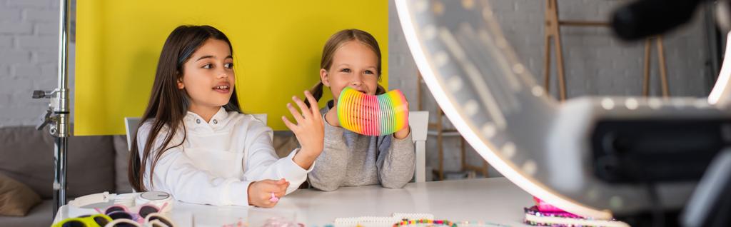 blogueiro morena falando perto de menina sorridente mostrando brinquedo espiral na frente da lâmpada círculo borrada, banner - Foto, Imagem