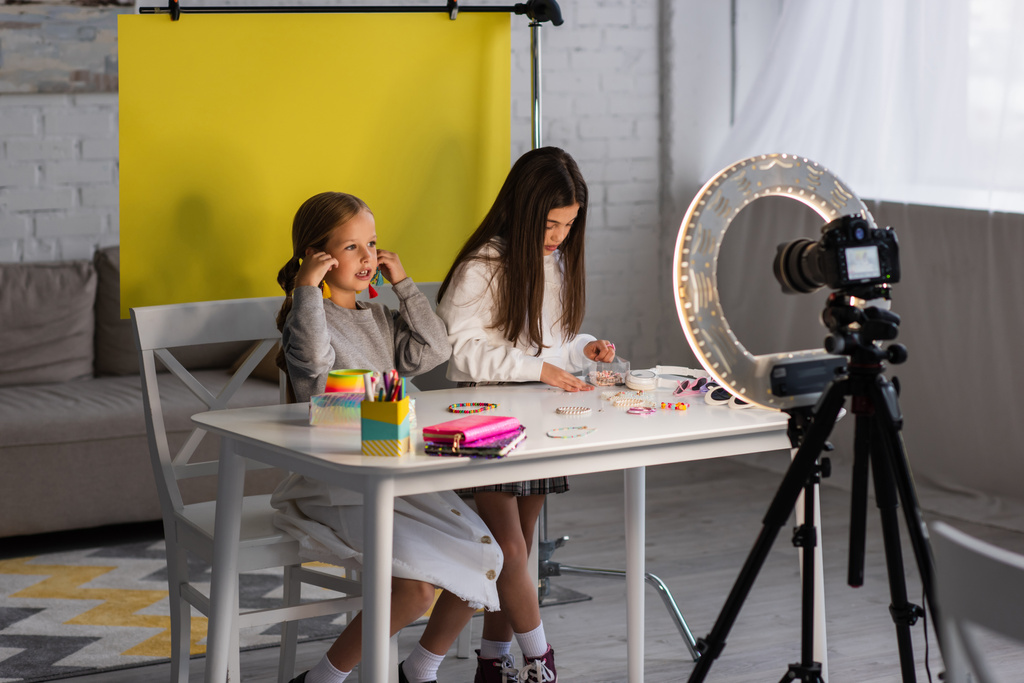 tiener video blogger praten in de buurt meisje en verschillende accessoires op tafel in de voorkant van digitale camera in ring licht - Foto, afbeelding