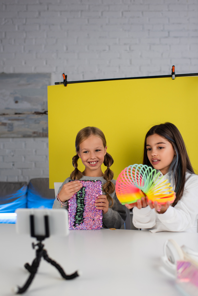 video bloggers met spiraalvormig speelgoed en glanzend copybook op gele achtergrond voor wazig smartphone op houder - Foto, afbeelding