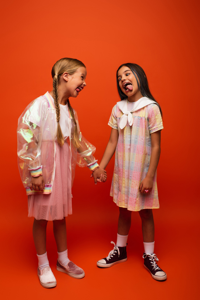 teljes hosszúságú vicces lányok fogják egymás kezét és kinyújtják a nyelvüket narancssárga alapon - Fotó, kép