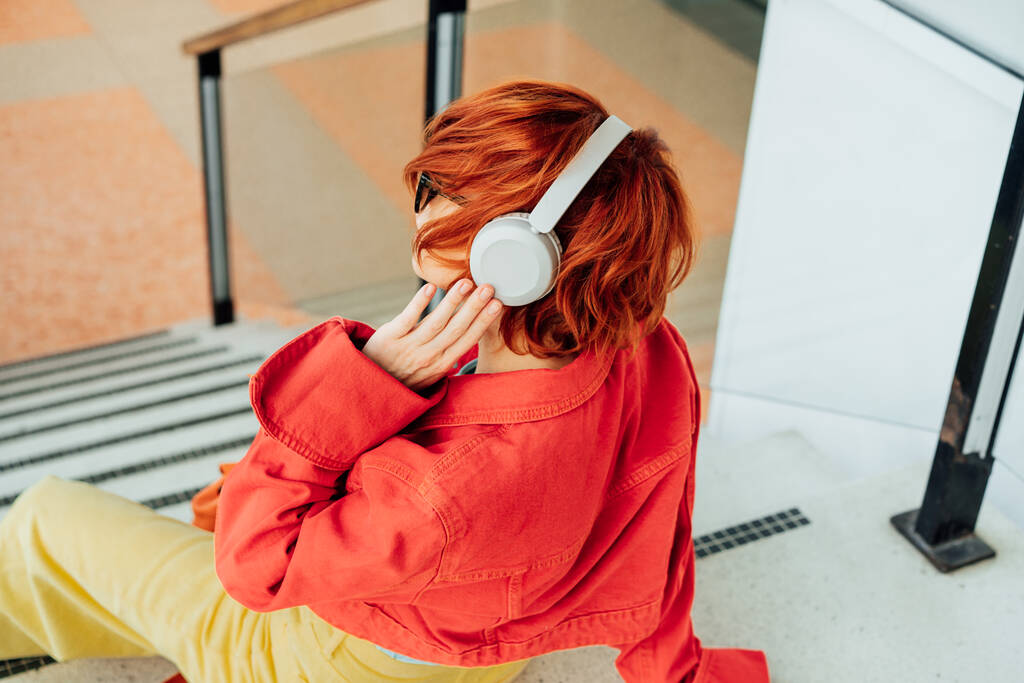Back view hipster giovane donna in abiti luminosi, occhiali da sole e cuffie wireless ascoltare musica mentre seduto sulle scale in luogo pubblico. Moda urbana di strada. Focus selettivo - Foto, immagini
