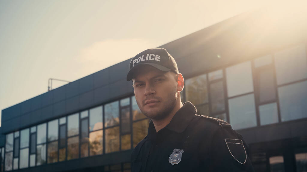 young policeman in cap with lettering and uniform looking at camera near building - Photo, Image