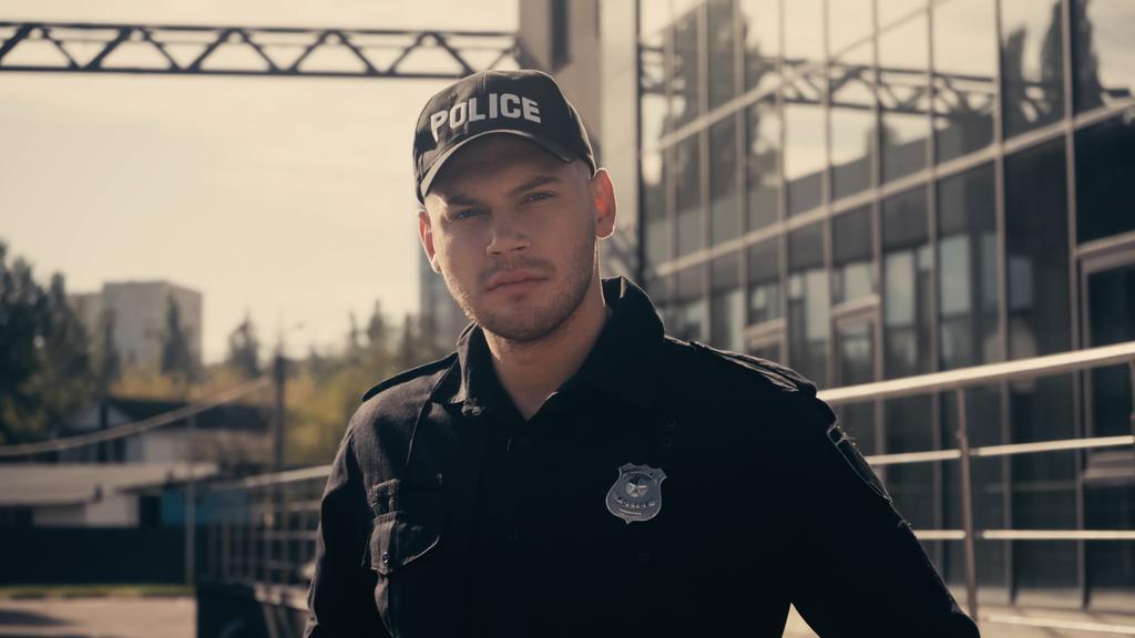 jeune policier en casquette avec lettrage et uniforme regardant la caméra à l'extérieur  - Photo, image