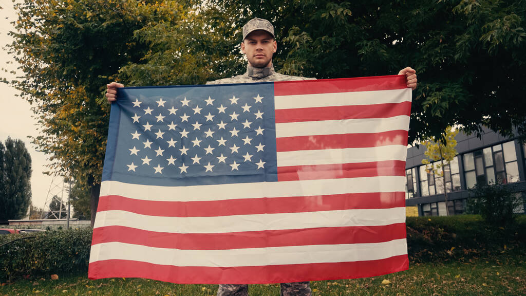 Soldado en uniforme militar y gorra con bandera americana mientras mira la cámara al aire libre  - Foto, Imagen