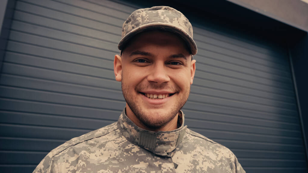 soldat joyeux en uniforme et casquette regardant la caméra près du bâtiment  - Photo, image