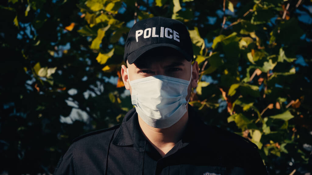 портрет молодого полицейского в форме и медицинской маске, стоящего рядом с деревом  - Фото, изображение