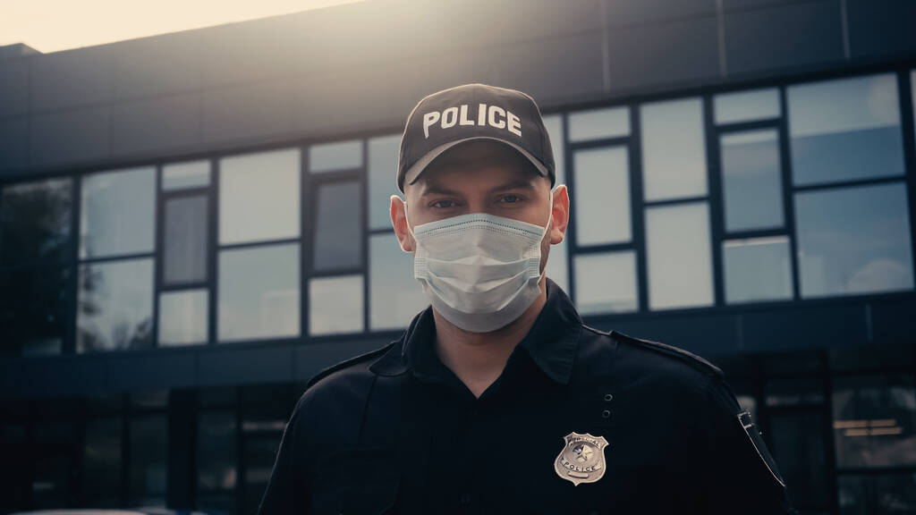 policier en masque médical et uniforme regardant la caméra près du bâtiment  - Photo, image