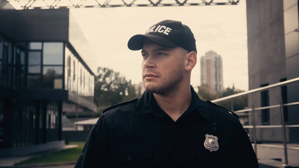 jeune policier debout en casquette et uniforme regardant loin tout en patrouillant la rue - Photo, image