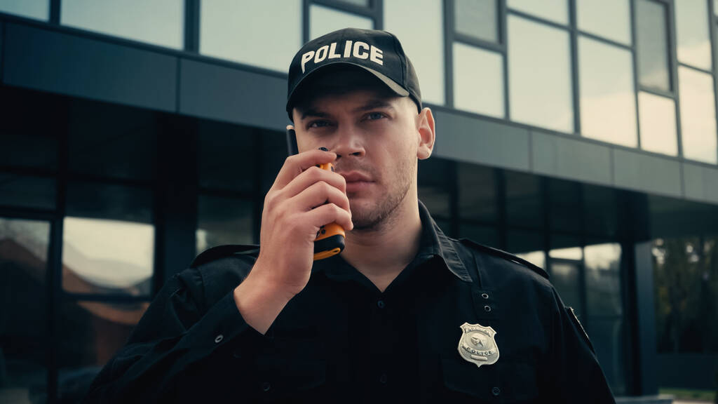 młody policjant w mundurze i czapce informujący przez krótkofalówkę na zewnątrz  - Zdjęcie, obraz