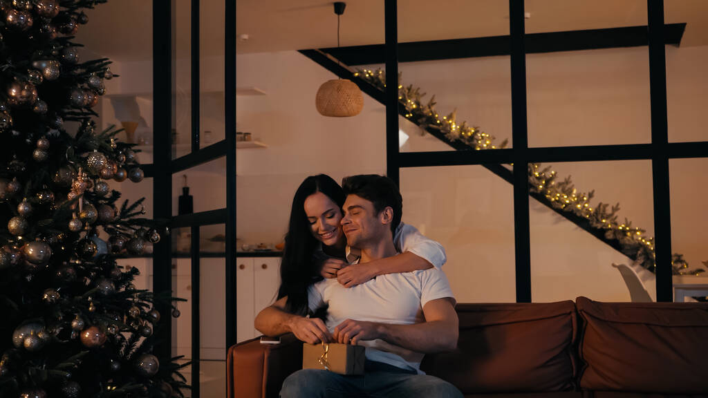 mulher feliz abraçando namorado presente de abertura enquanto sentado no sofá perto de smartphone e árvore de Natal - Foto, Imagem