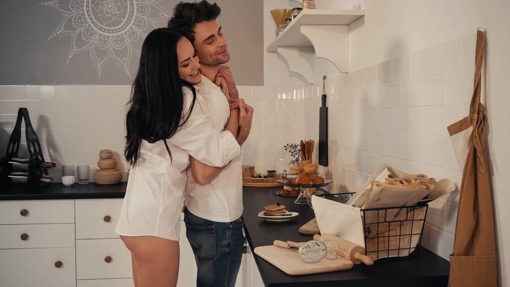 feliz jovem mulher em camisa branca abraçando namorado alegre na cozinha  - Foto, Imagem