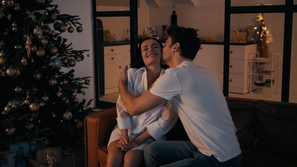Mann küsst Wange einer glücklichen Frau, die auf Couch in der Nähe des Weihnachtsbaums sitzt - Foto, Bild