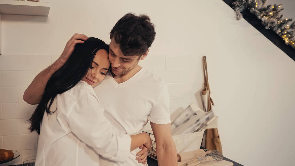 bruna giovane donna in camicia bianca abbracciare fidanzato accarezzando i capelli in cucina  - Foto, immagini