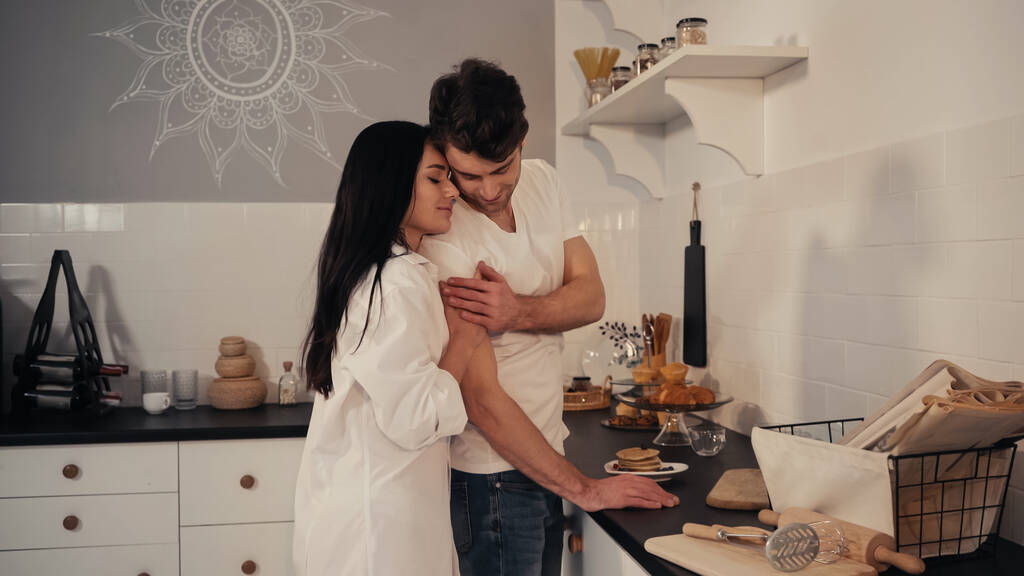 брюнетка и чувственная женщина в белой рубашке обнимают счастливого мужчину на современной кухне  - Фото, изображение