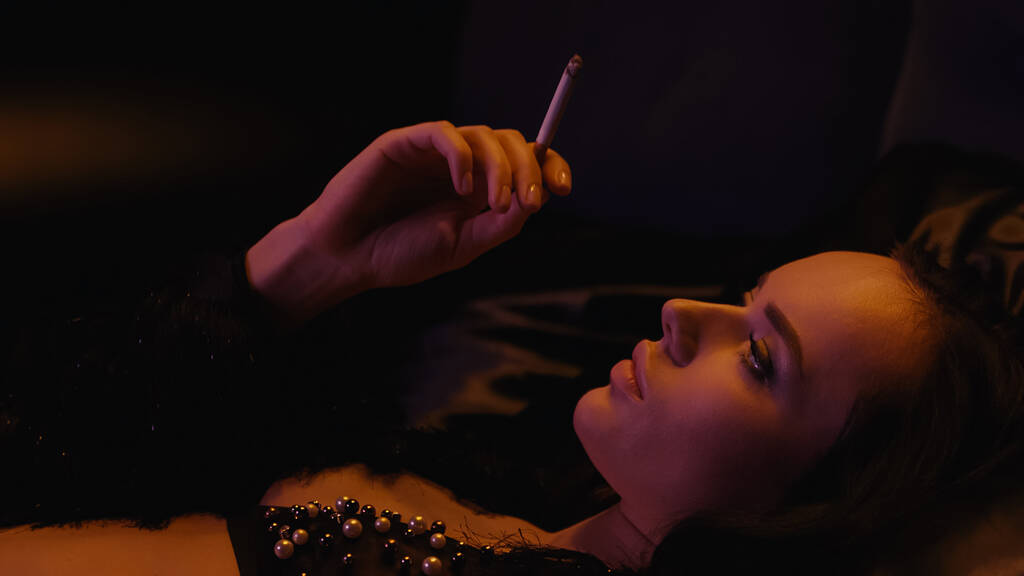 Μελαχρινή γυναίκα με σουτιέν κρατώντας τσιγάρο και ξαπλωμένη σε μαύρο φόντο  - Φωτογραφία, εικόνα
