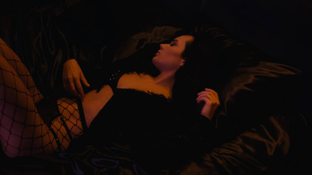 Вид сбоку чувственной женщины в нижнем белье и колготках-фишнетах, лежащей на кровати  - Фото, изображение