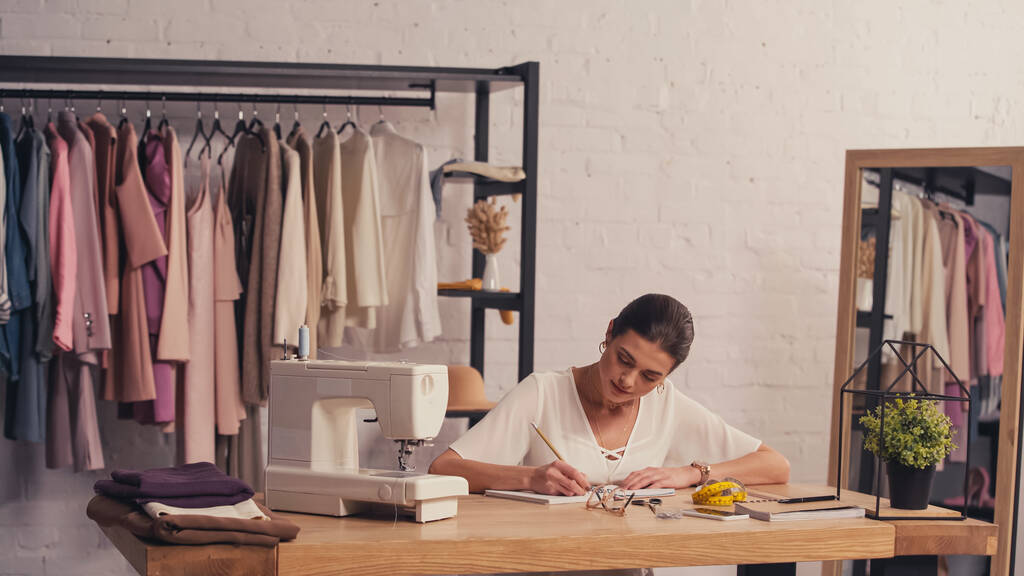 Designer écrivant sur carnet près de tissu et machine à coudre en atelier  - Photo, image