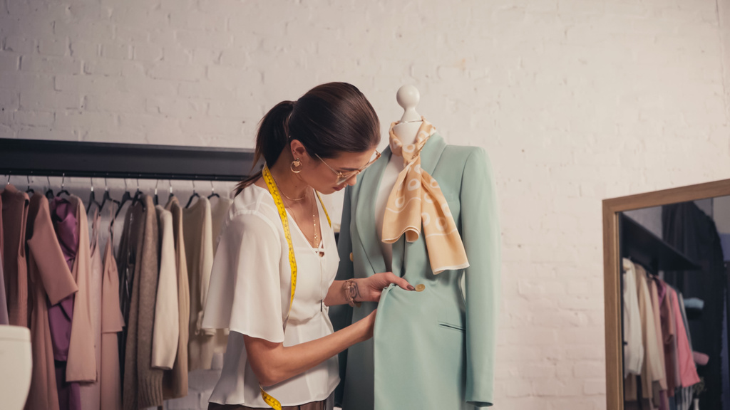 Brunette ontwerper met meetlint werken met blazer op mannequin in werkplaats  - Foto, afbeelding