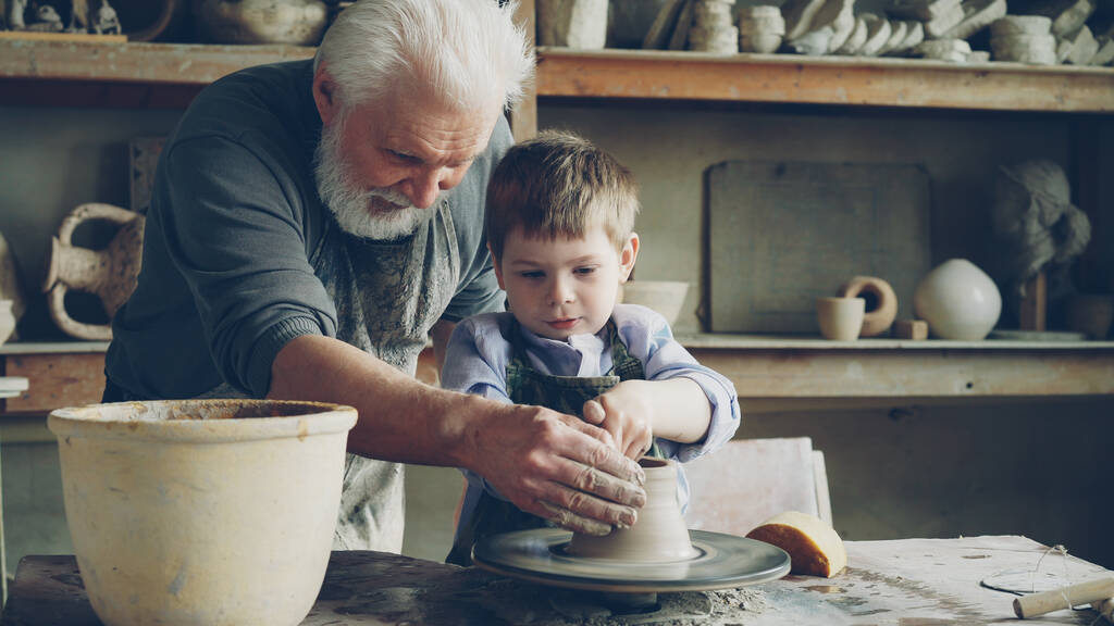 Skoncentrowany mały chłopiec uczy się pracować z gliny na profesjonalnych rzucanie koła w klasy ceramiki w tradycyjnych warsztatach. Pomaga mu jego nauczyciel starszy człowiek doświadczony. - Zdjęcie, obraz