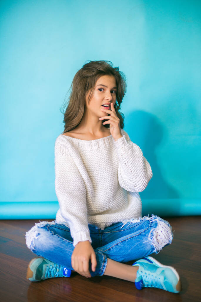 A cute teenage girl with long dark hair in trendy jeans and a white knitted sweater on a blue background in the studio. Fashion for teenagers - Photo, Image