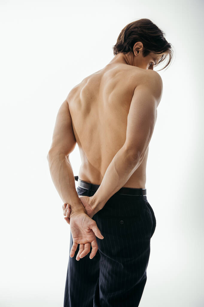vista posterior del hombre musculoso y sin camisa en pantalones negros posando con las manos detrás de la espalda en blanco - Foto, imagen