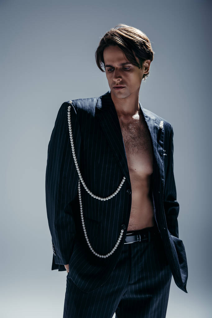 стильний чоловік з волохатими грудьми і костюм з перловим ланцюжком позує руками в кишенях на темно-сірому
 - Фото, зображення