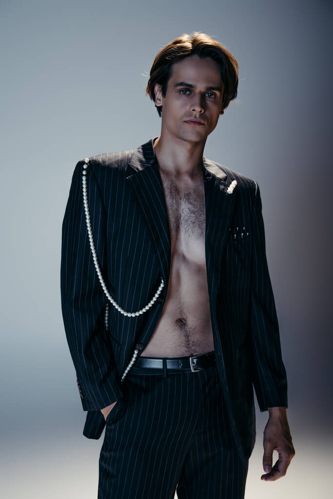hombre elegante con el pecho peludo y traje de moda con cadena de perlas posando con la mano en el bolsillo en gris oscuro - Foto, Imagen