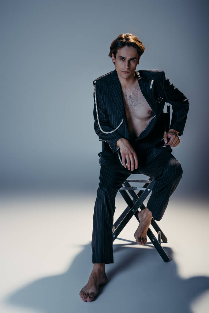 volle Länge des stilvollen Mannes mit behaarter Brust und trendigem Anzug mit Perlenkette sitzt auf einem Stuhl auf dunkelgrau  - Foto, Bild