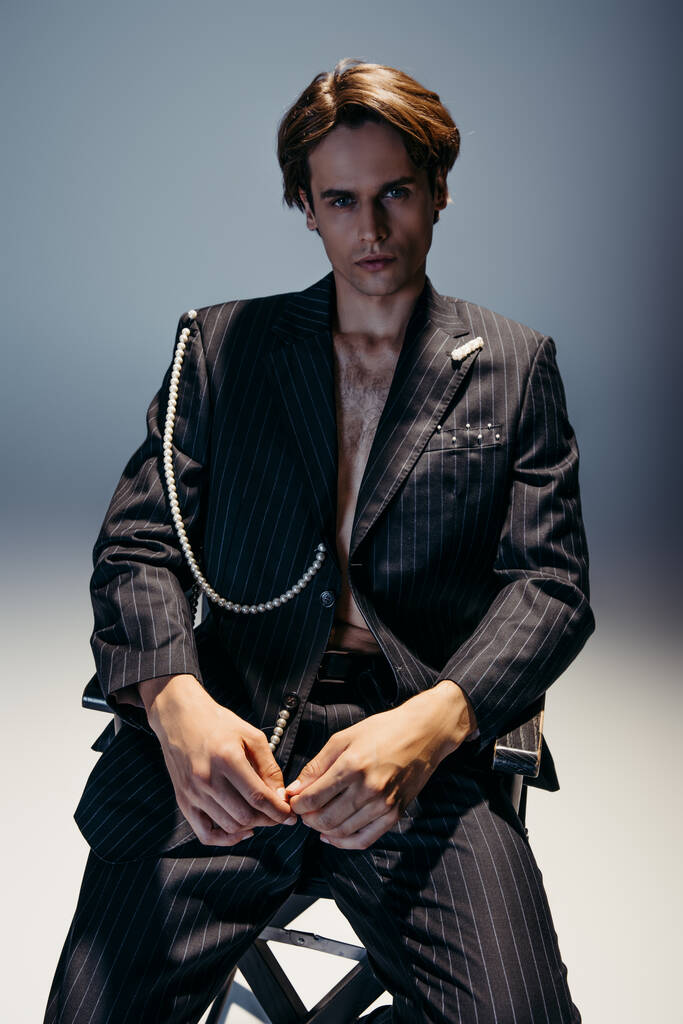 stylischer Mann im trendigen Anzug mit Perlenkette sitzt auf einem Feldstuhl auf dunkelgrau  - Foto, Bild