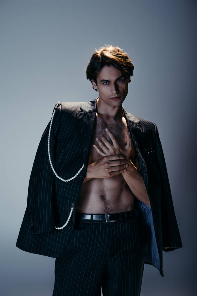стильный мужчина в модном костюме с жемчужной цепью и брюками, смотрящий в камеру на темно-сером  - Фото, изображение