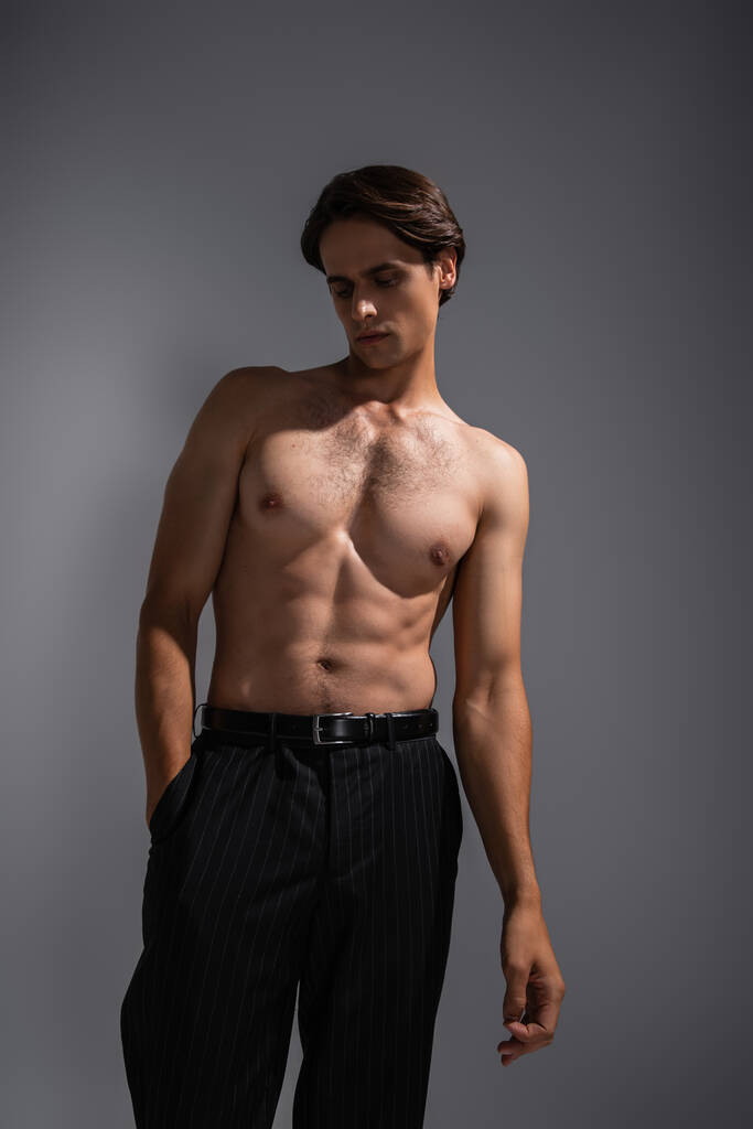 hombre guapo con cuerpo musculoso posando con la mano en el bolsillo de pantalones negros en gris - Foto, imagen
