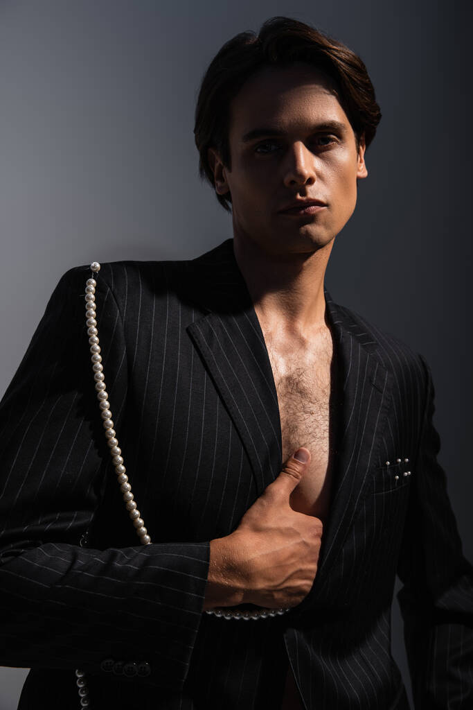 άντρας με μοντέρνο κοστούμι με πέρλες που κοιτάζει την κάμερα ενώ ποζάρει σε σκούρο γκρι   - Φωτογραφία, εικόνα