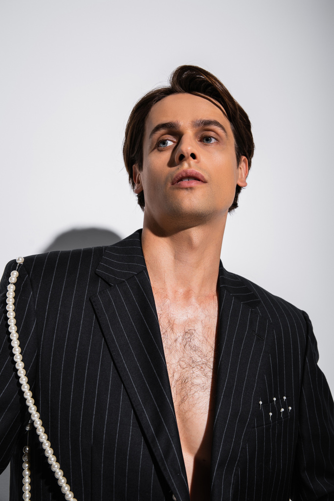 morena hombre en chaqueta de moda con rayas y perlas posando mientras mira hacia otro lado en gris - Foto, Imagen