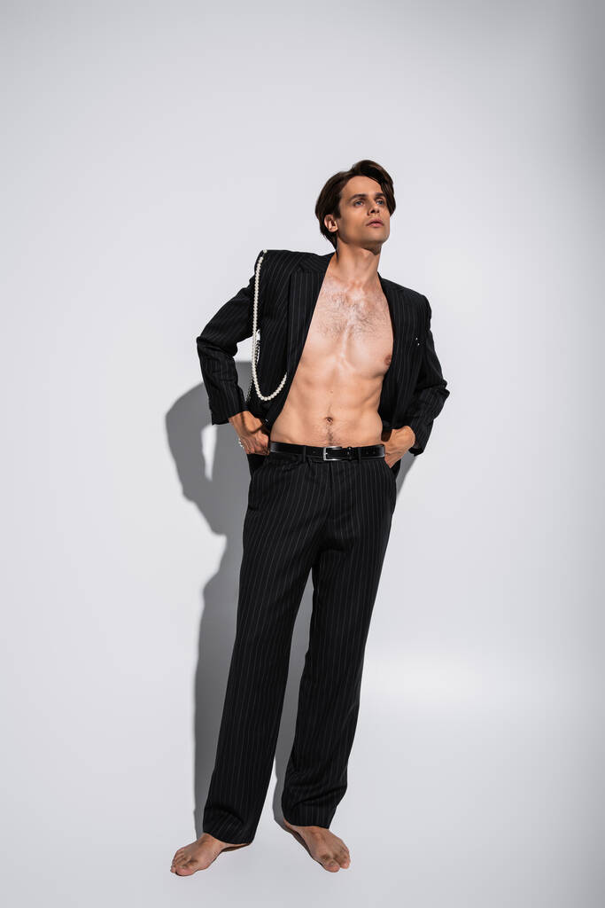 full length of pensive man σε μοντέρνο κοστούμι με πέρλες που στέκεται με τα χέρια στις τσέπες στο γκρι  - Φωτογραφία, εικόνα