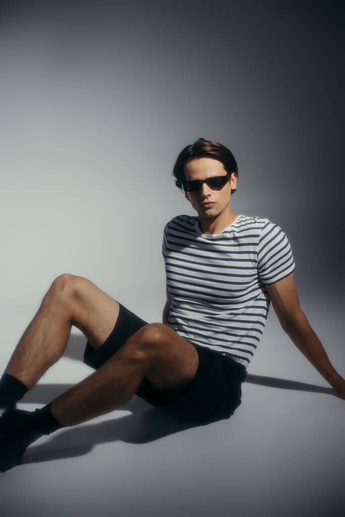 Brunette man in striped t-shirt and shorts sitting on grey background with lighting  - Photo, Image