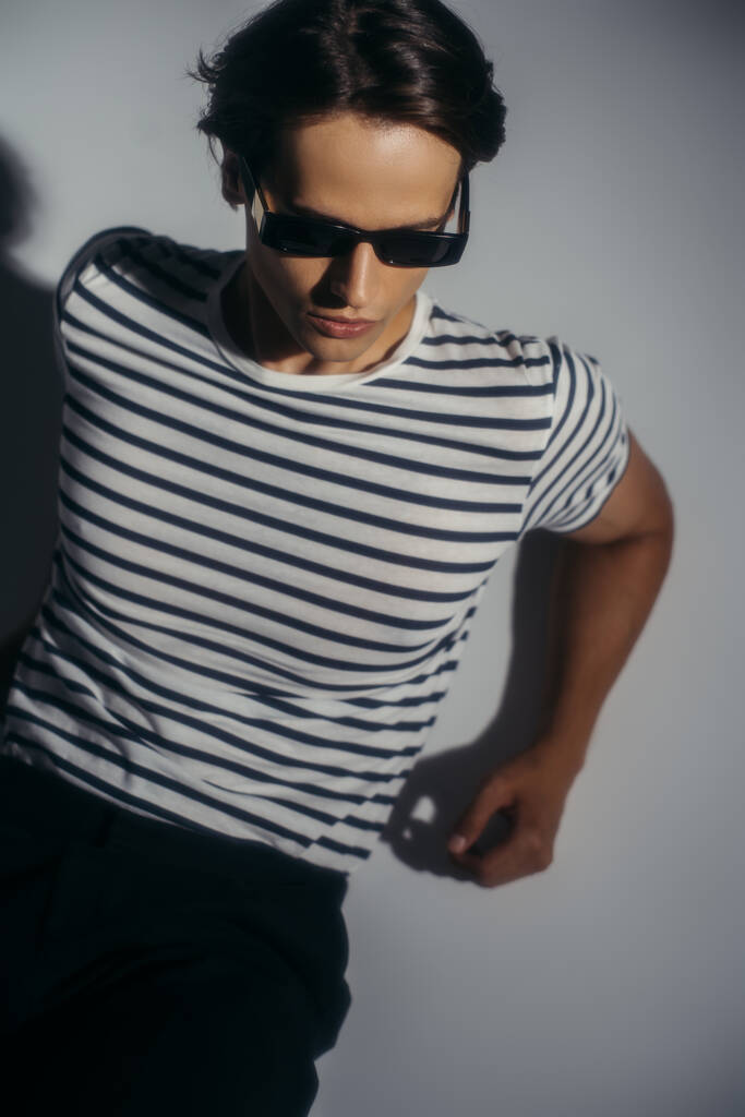Vista ad alto angolo del modello in t-shirt a righe e occhiali da sole su sfondo grigio con illuminazione  - Foto, immagini