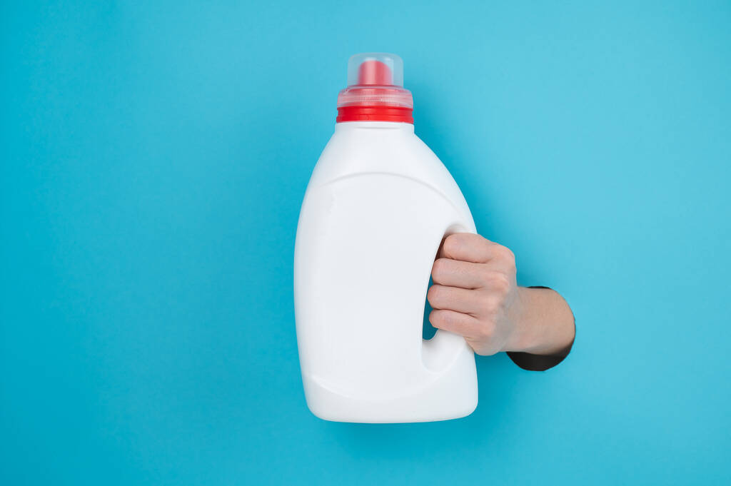 Una mujer sostiene una botella blanca de detergente. Mujeres mano sobresaliendo de fondo de papel azul - Foto, Imagen