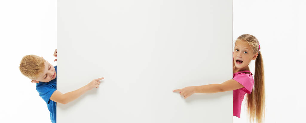 Стильный мальчик и девочка весело смеются с широко раскрытым ртом в повседневной одежде, выглядывая из-за белой стены. Копирование текста - Фото, изображение
