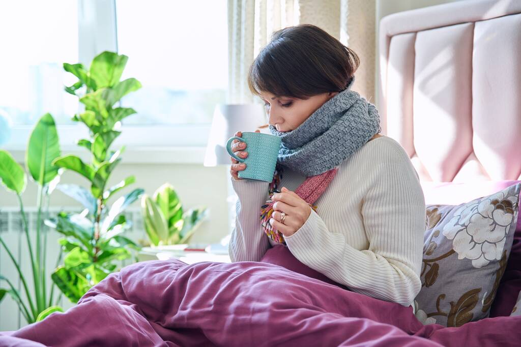 Mujer de mediana edad sentada en la cama bajo la manta calentándose con bufanda y bebida caliente en taza, estilo de vida casero en frío otoño invierno temporada - Foto, imagen