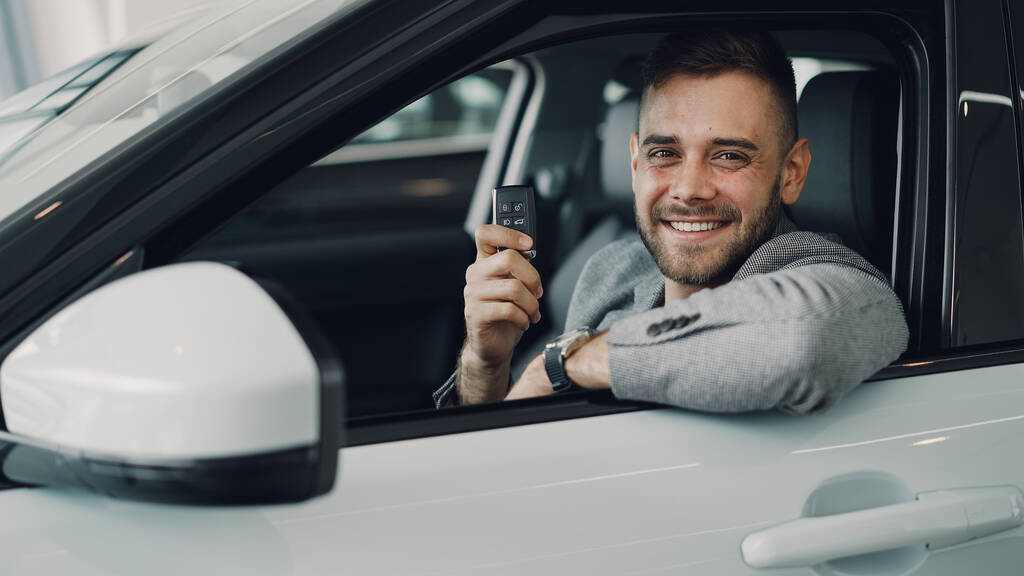 Közelkép portréja szép szakállas fickó új gépkocsi-tulajdonosnak benn gyönyörű automobile üzem kulcstartó, és mosolyogva nézi a kamera. Szállítás és az emberek koncepció. - Fotó, kép