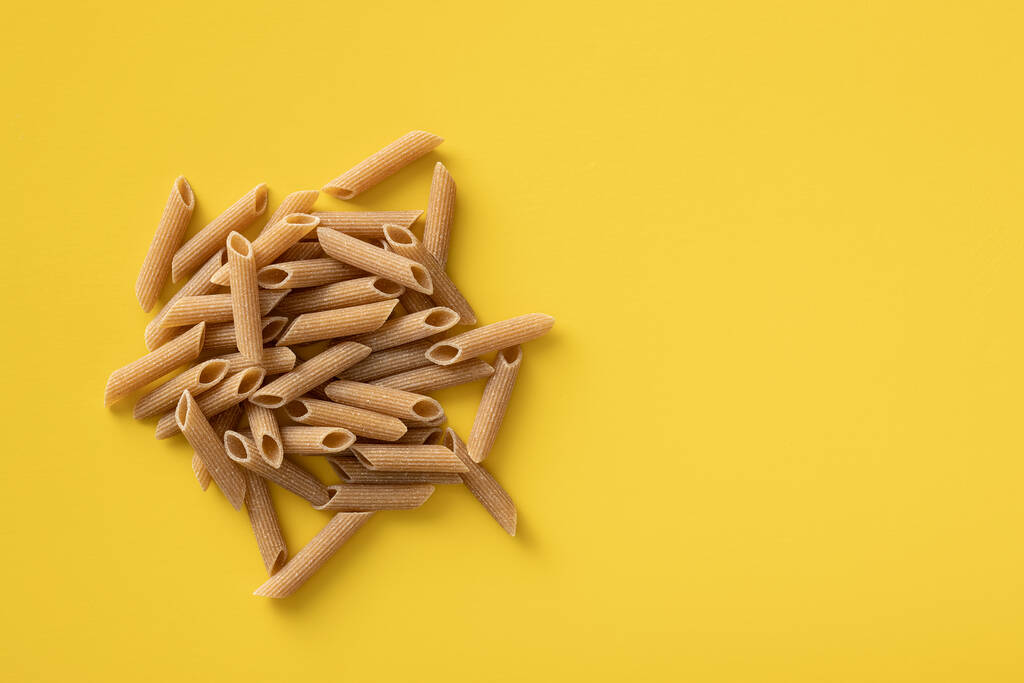 The uncooked whole grain pasta. Raw penne pasta on yellow background. Top view. - Photo, Image