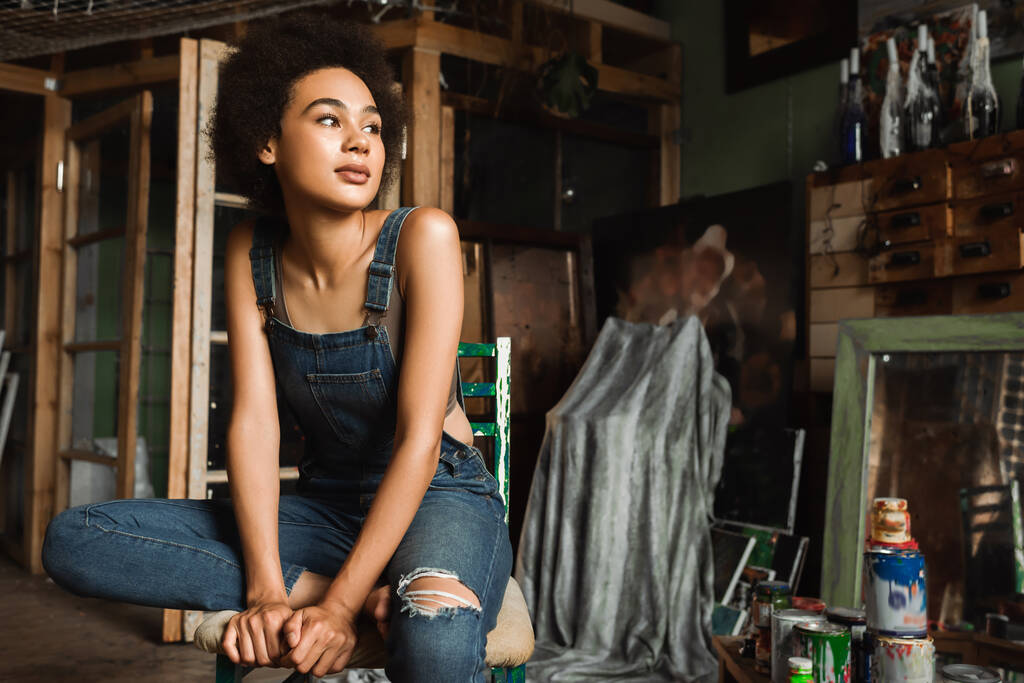 joven mujer afroamericana en overol denim mirando hacia otro lado en taller de arte cerca de latas con pintura - Foto, imagen