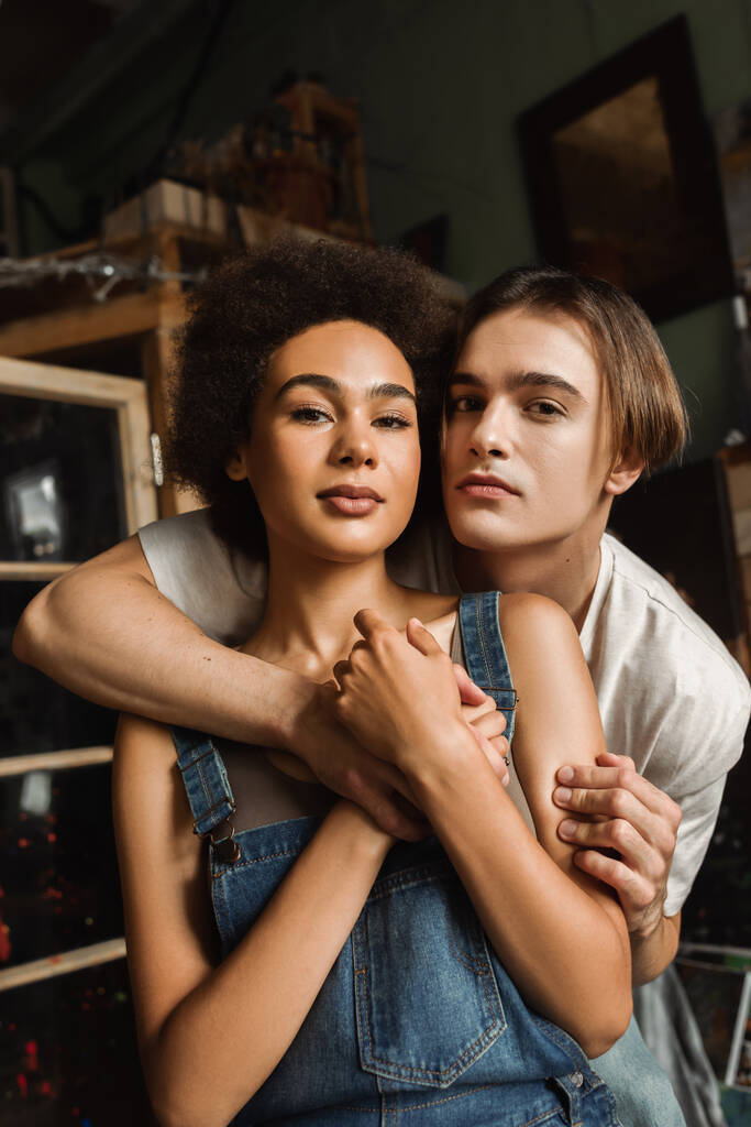 νεαρό διαφυλετικό ζευγάρι αγκαλιάζει και κοιτάζοντας κάμερα στο στούντιο τέχνης - Φωτογραφία, εικόνα