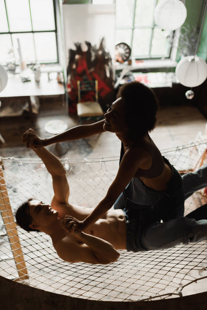 веселый мужчина без рубашки, держась за руки с африканской американской подружкой, веселясь в гамаке над мастерской - Фото, изображение
