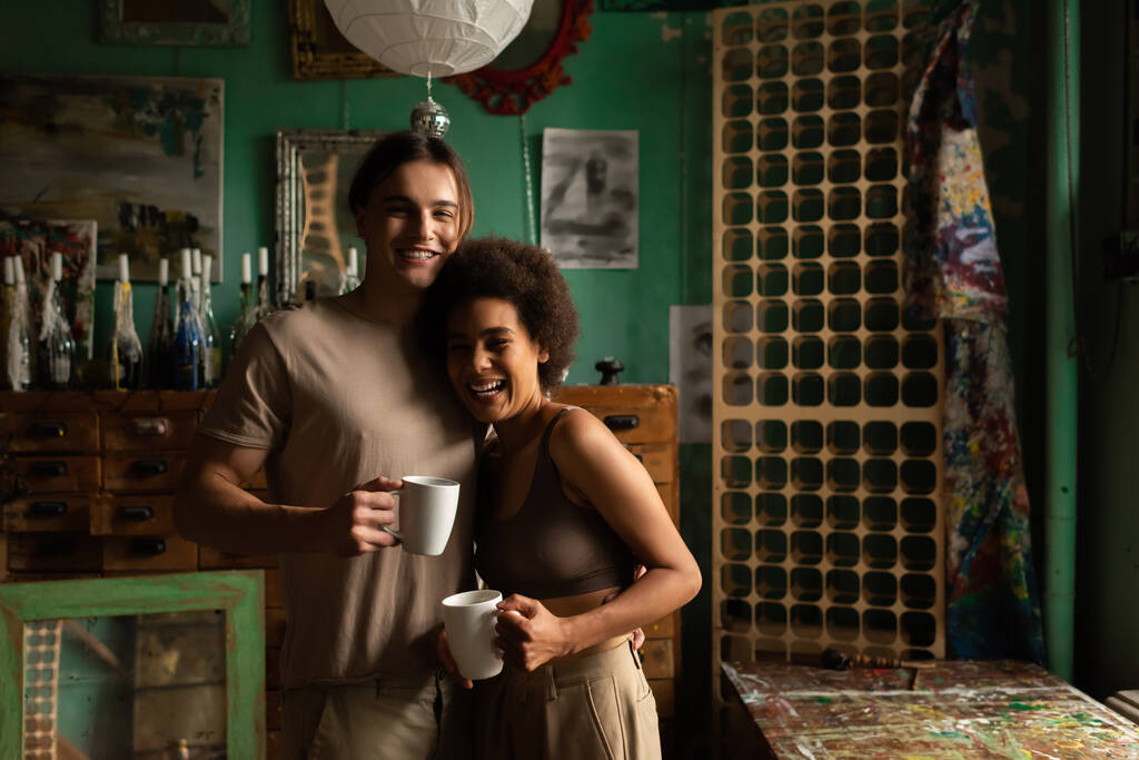 счастливая межрасовая пара с чашками чая глядя на камеру в студии с винтажным декором - Фото, изображение