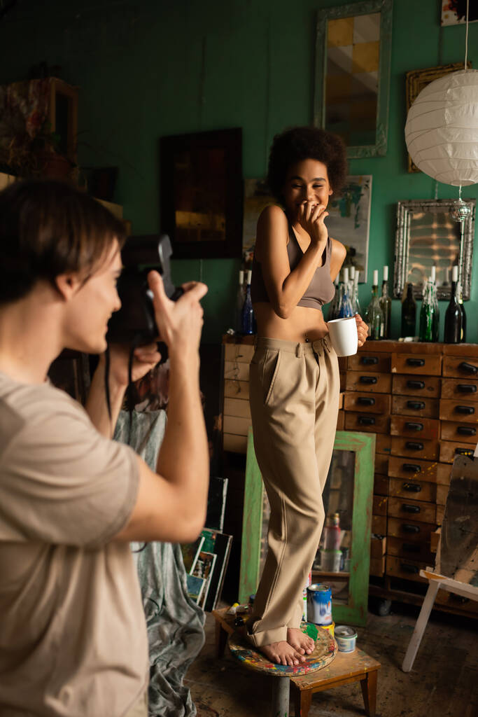 zamazany mężczyzna z klasycznym aparatem robienie zdjęć afrykańskiej dziewczyny amerykańskiej stojącej na stołku z filiżanką herbaty - Zdjęcie, obraz